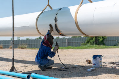 Worker in protective suit and mask crouching and welding pipe of water tower at construction site.