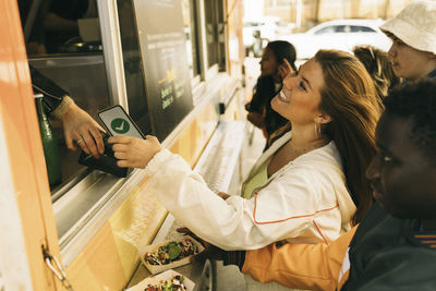 Smiling young woman paying via tap to pay while buying food from concession stand