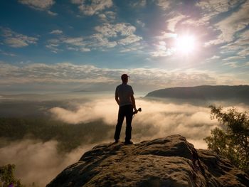 Tall photographer with baseball cap and tripod with camera in hands on rocky view and watching down 