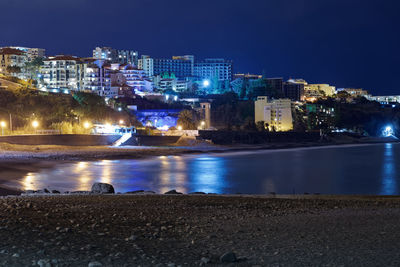 Illuminated buildings by sea against sky at night