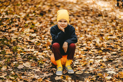 A boy a teen child in a bright yellow hat sits on a pumpkin in an autumn forest in nature outdoors