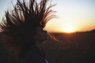 Side view of woman with eyes closed tossing hair while standing against sky during sunset
