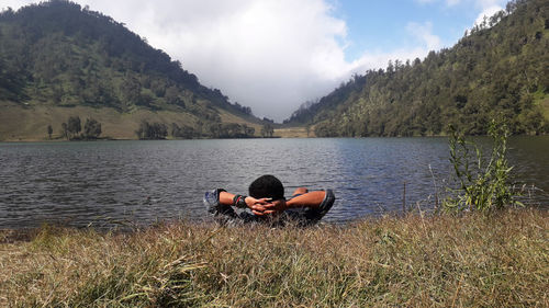 Man lying on grass by lake against mountains and sky