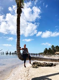 Portrait of woman sitting on tree trunk at beach against blue sky