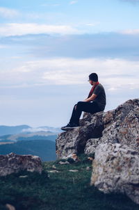 Man sitting on rock looking at mountain against sky