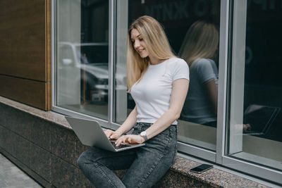 Young woman using laptop while sitting by window outdoors