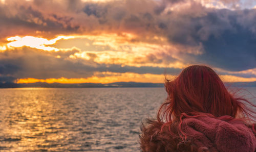 Rear view of woman with redhead sitting at seashore against dramatic sky