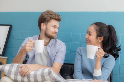 Friends holding coffee cups while looking each other at home