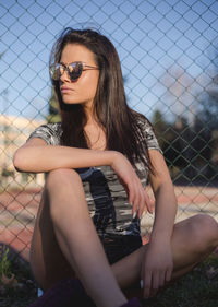 Young woman wearing sunglasses while sitting by chainlink fence
