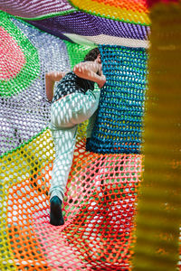 Adorable cheerful little boy with dark hair in casual clothes playing on colorful knitted trampoline in outdoor entertainment center