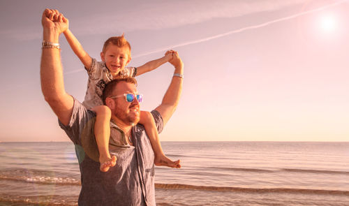 Man piggybacking son while standing at beach against sky during sunset