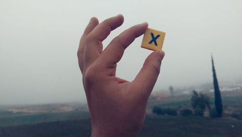 Cropped hand holding cross block against sky