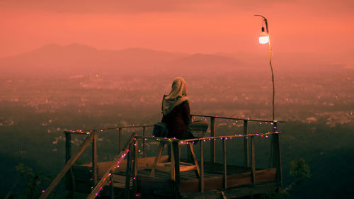 Rear view of woman sitting on rooftop by railing against sky during sunset