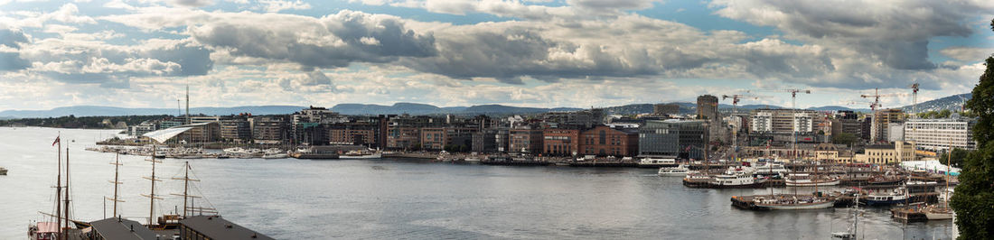 Panoramic view of oslo port in city by sea against cloudy sky