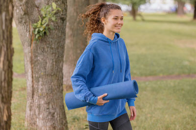 Smiling woman holding exercise mat standing at park