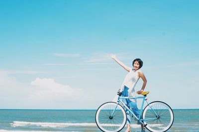 Portrait of smiling young woman standing by bicycle at beach against sky