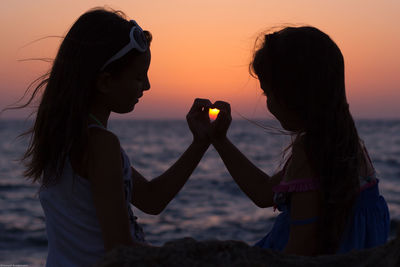 Girls making heart shape with hands while sitting at sea shore during sunset