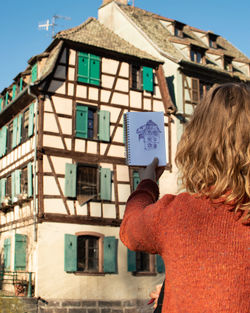 Rear view of woman standing with drawing in book against building in city