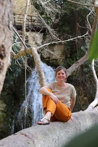 Portrait of smiling young woman sitting on tree trunk in forest