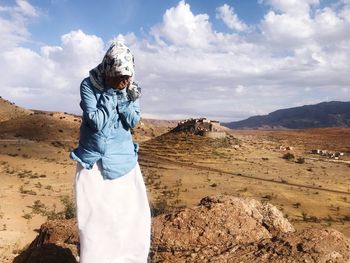 Young woman covering face while standing on cliff against cloudy sky