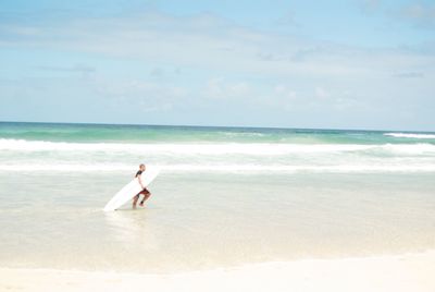 Side view of man with surfboard while walking in sea against sky