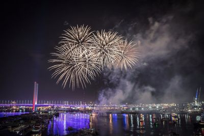 Firework display over sea and bridge in city against sky at night