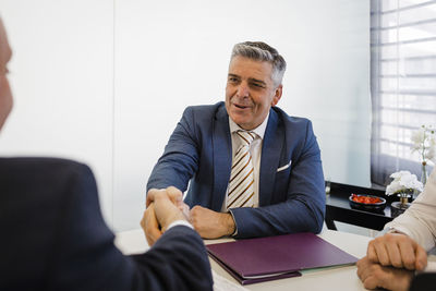 Mature businessman shaking hands with colleague sitting at desk