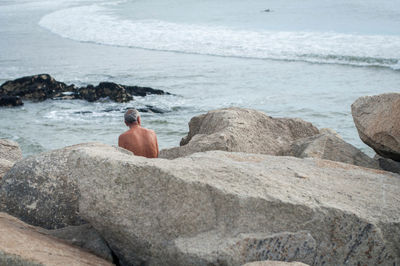 Shirtless man sitting by rocks at beach