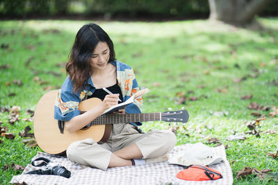 Young woman writing in diary while holding guitar while sitting at lawn