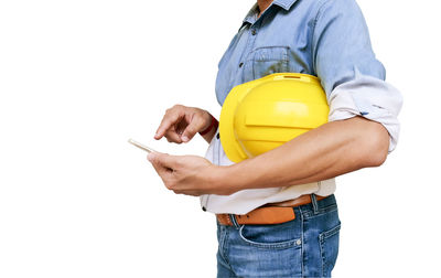 Midsection of architect holding hardhat while using smart phone against white background