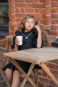 Portrait of beautiful young woman drinking coffee while sitting on chair against brick wall