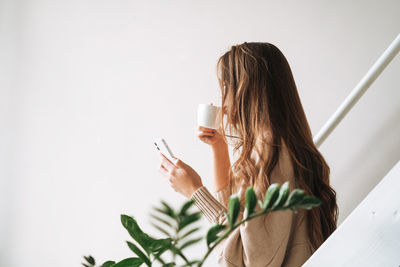 Young woman with brunette long hair in cardigan with cup of tea in hands using mobile phone iat home