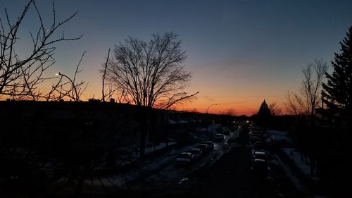 Silhouette bare trees by road against sky at sunset