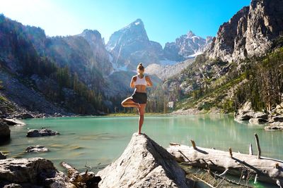 Rear view of young woman exercising on rock by lake against sky