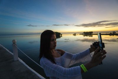 Woman photographing sea against sky during sunset