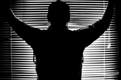 Rear view of silhouette man standing by window blinds at home