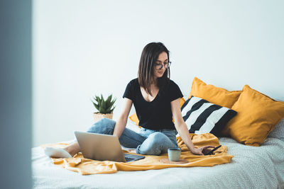 Female entrepreneur sitting on bed with laptop computer and checking smartphone while working home 