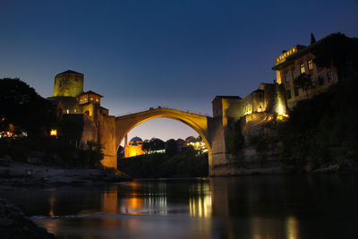 Bridge over river by illuminated buildings against clear sky at night