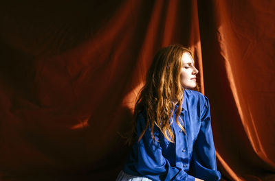 Young woman with eyes closed sitting in front of red cloth in sunlight