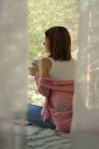 Beauty young woman looking on window from cozy home and drink coffee or tea