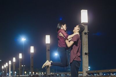 Side view of man holding girlfriend against at night