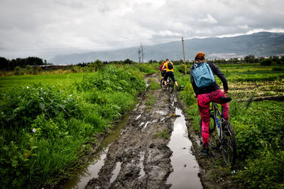 Rear view of people riding bicycle on dirt road against sky