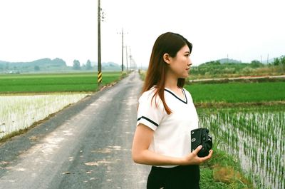 Beautiful woman holding camera while standing on road amidst rice paddy