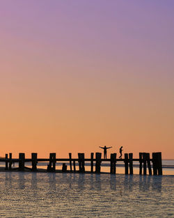 Silhouette wooden posts in sea against orange sky