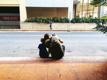 Rear view of father and son photographing while sitting on footpath by street