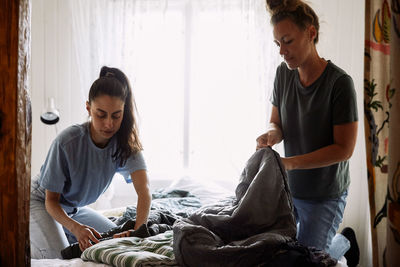 Female friends folding blankets while kneeling on bed against window in cottage