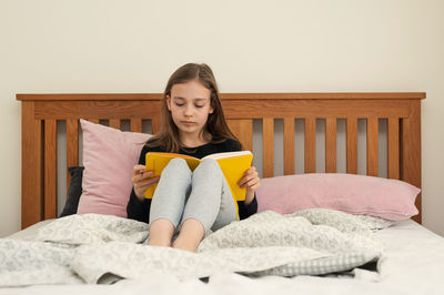 Young school age girl sitting on bed in bedroom and reading from yellow textbook. school homework.