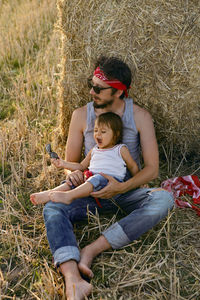 Father and son in t-shirts sitting next to a haystack on a sloping field during sunset