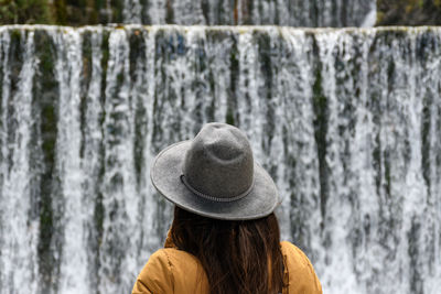 Rear view of woman wearing warm clothes and hat, standing in front of waterfall.