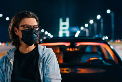 Portrait of man with medical mask standing by car in illuminated city at night